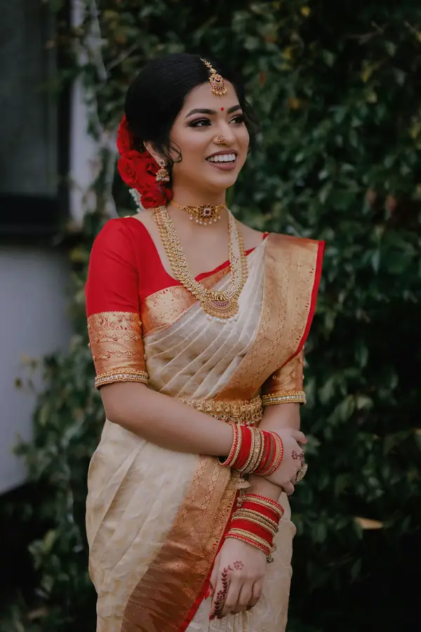 A bride in a cream and red silk saree standing in a garden