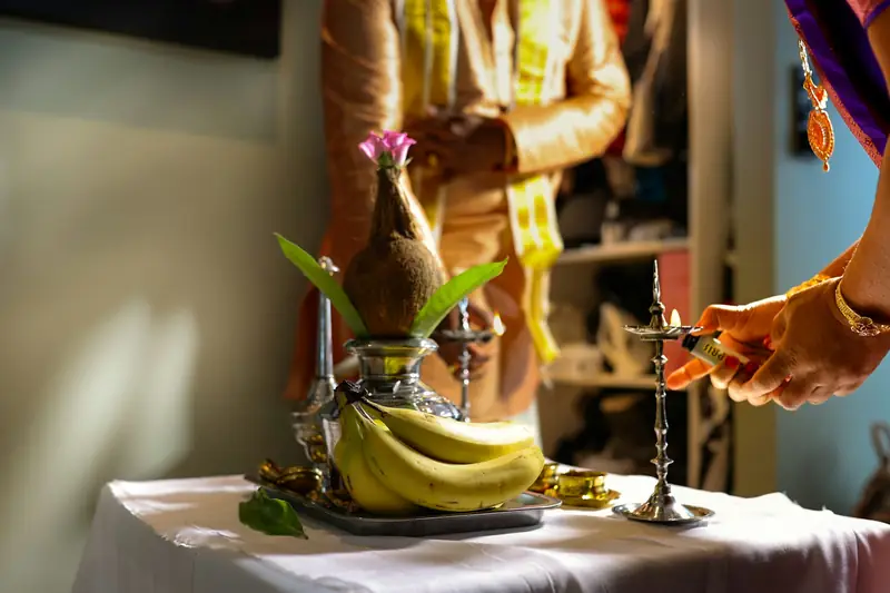 A kalasam with coconut and mango leaves, bananas, and a brass lamp being lit