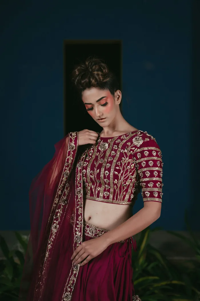 Woman in a maroon blouse covered in gold maggam work, with a matching dupatta, against a teal wall