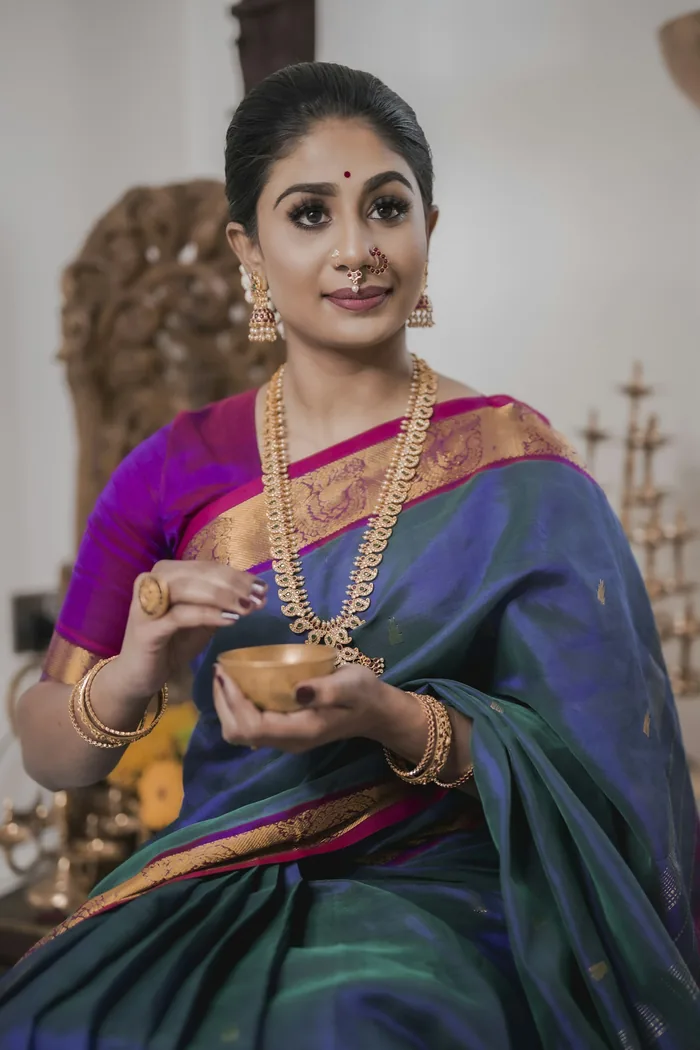 Woman in a peacock blue silk saree with a gold border and a purple blouse, holding a small brass bowl